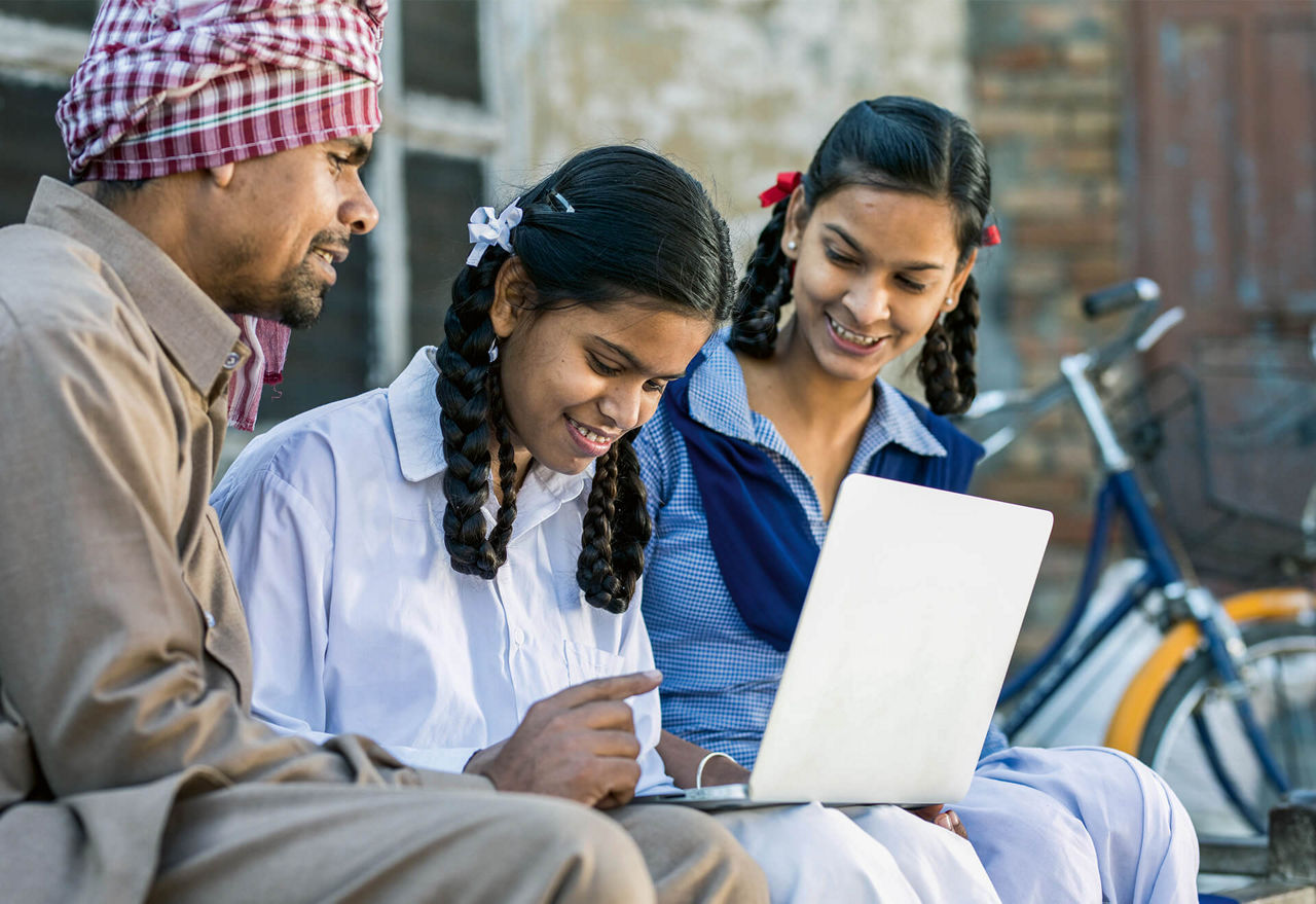 Two young women and a man are sitting outside looking at a laptop together.