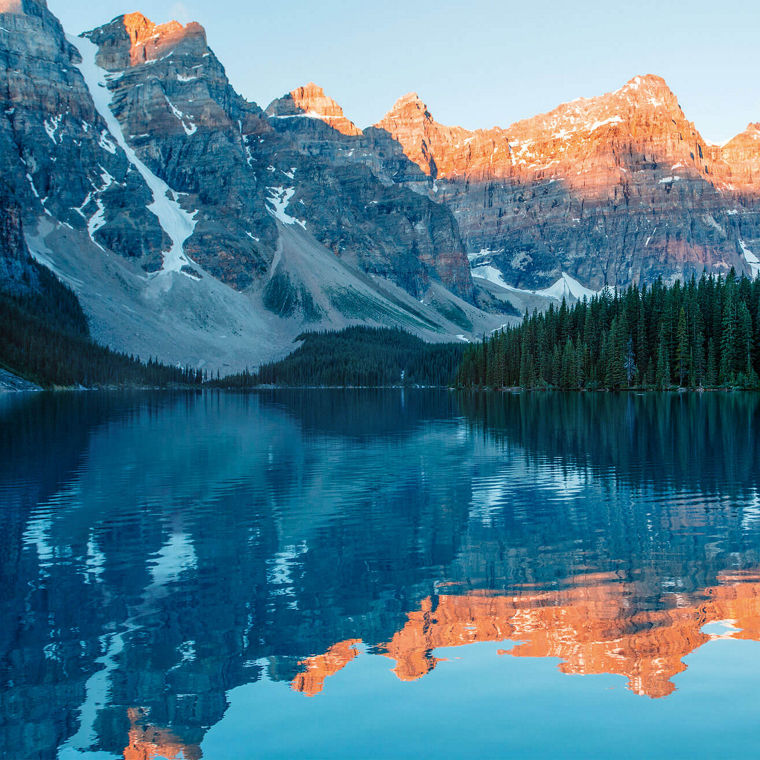 Ein ruhiger kanadischer See in einem Nadelwald, dahinter Berggipfel im Sonnenschein.