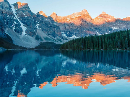Ein ruhiger kanadischer See in einem Nadelwald, dahinter Berggipfel im Sonnenschein.