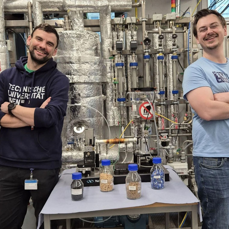 Zwei junge Wissenschaftler stehen mit verschränkten Armen neben Stoffproben. Sie Lachen in die Kamera.