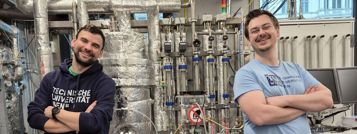 Zwei junge Wissenschaftler stehen mit verschränkten Armen neben Stoffproben. Sie Lachen in die Kamera.