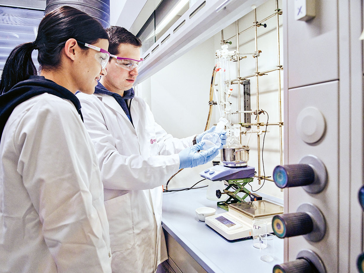 Two lab technicians in white coats are collaborating on an experiment at a laboratory workstation.