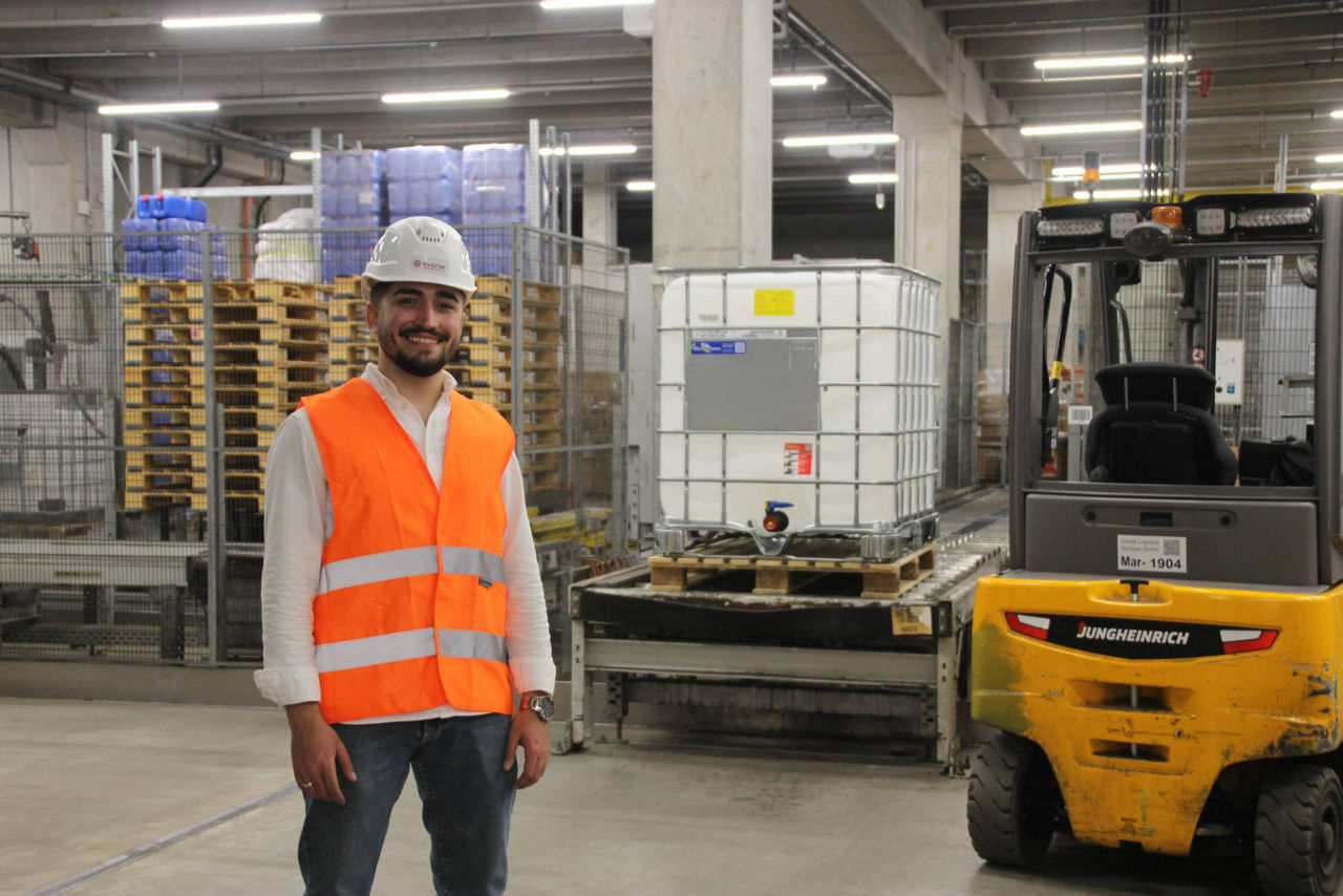 A worker in an orange safety vest stands in a warehouse with pallets and a forklift in the background.