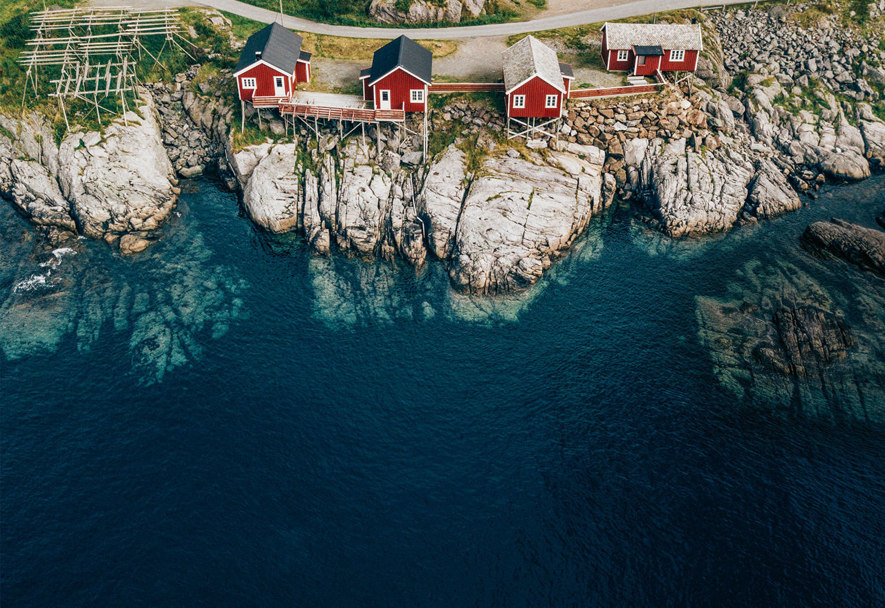 Rot gestrichene Häuser stehen, zum Teil auf Holzbalken gestützt, auf einer Felskante eines Fjordes.