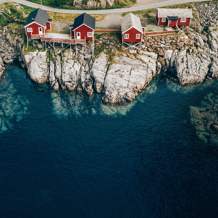 Rote Holzhäuser stehen zum Teil auf hohen Stelzen an der felsigen Klippe eines Fjords.