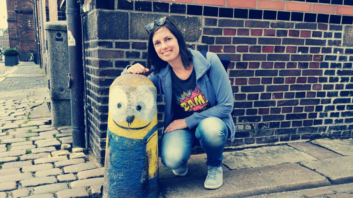 A person kneels next to a colorful minion-themed bollard on a cobblestone street.