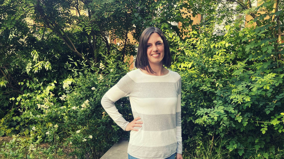 A person stands outdoors in front of lush greenery, wearing a striped long-sleeve shirt and jeans.