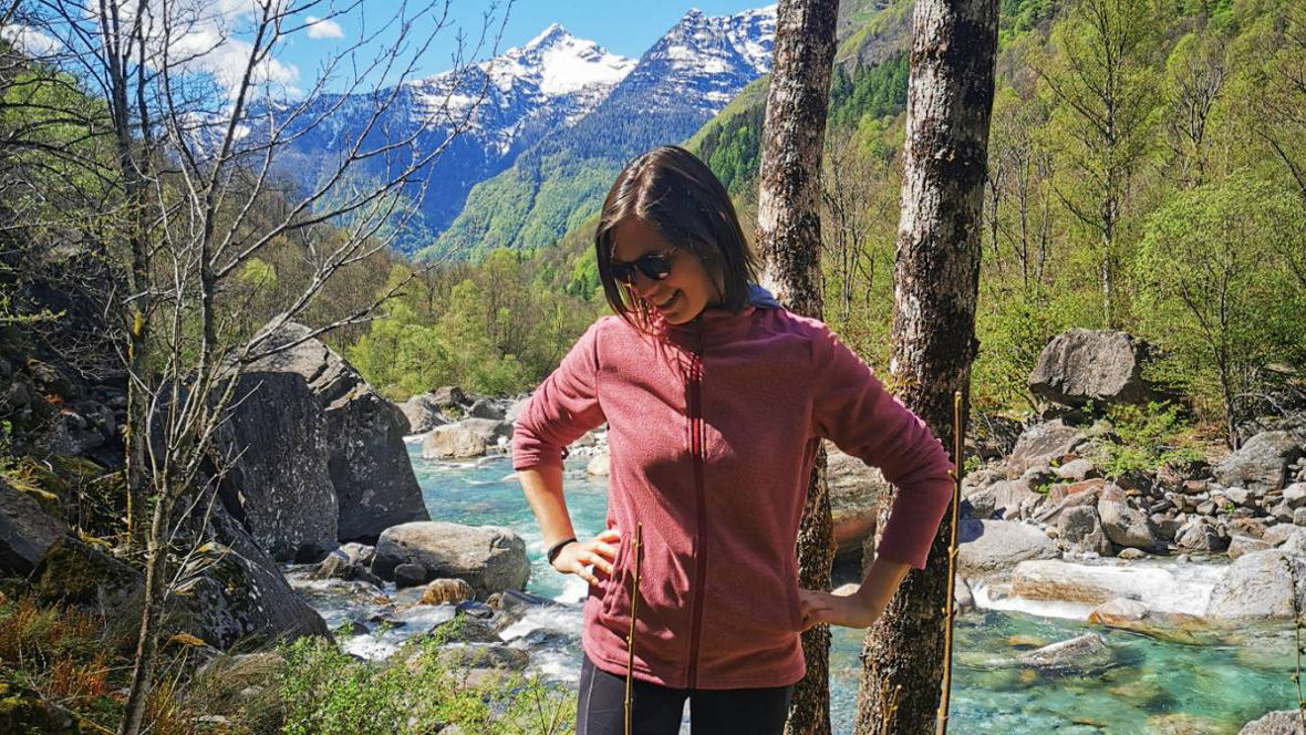 A person stands confidently by a river surrounded by lush greenery and mountains under a blue sky.