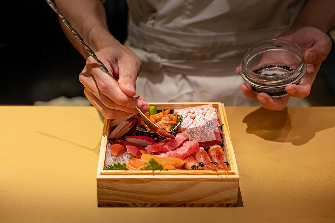 A man's hand holds a fine brush and paints carefully arranged pieces of fish with it.