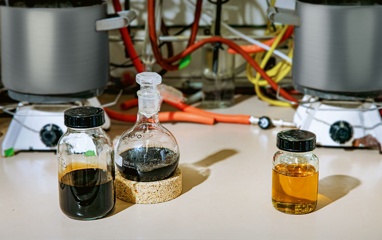 Auf einem Tisch stehen zwei verschlossene Glasbehälter mit tiefbrauner Flüssigkeit, daneben einer mit einer hellbraun-transparenten. Im Hintergrund sind Schläuche zu sehen.