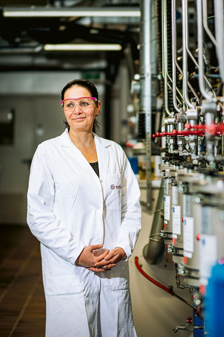 Annegret Terheiden steht neben einer Reihe von Leitungen. Sie trägt einen laborkittel und eine Schutzbrille.