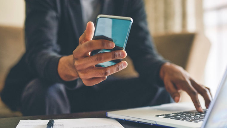 Man sitting on sofa checking laptop and smartphone with papers containing information in front of him.