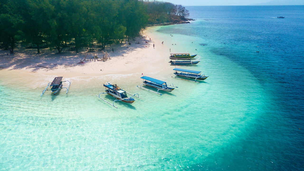 Excursion boats are moored on a white beach in crystal-clear water. There are only a few people on the beach, behind which a forest begins.