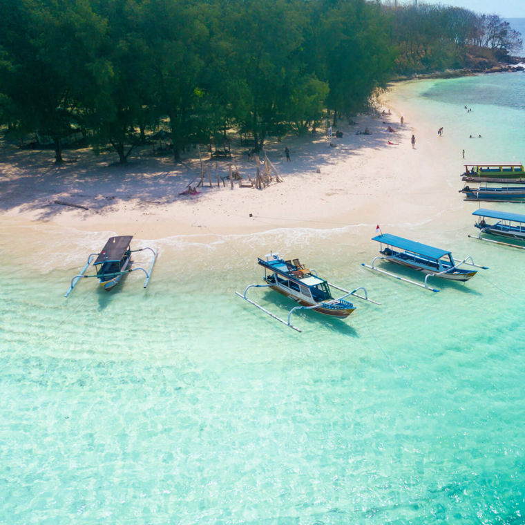 Excursion boats are moored on a white beach in crystal-clear water. There are only a few people on the beach, behind which a forest begins.