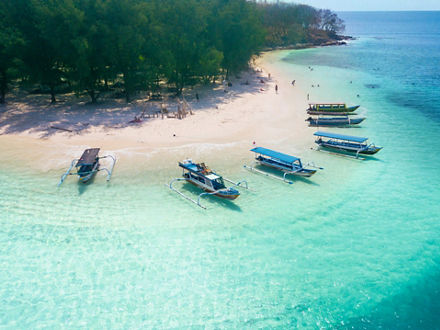 Excursion boats are moored on a white beach in crystal-clear water. There are only a few people on the beach, behind which a forest begins.