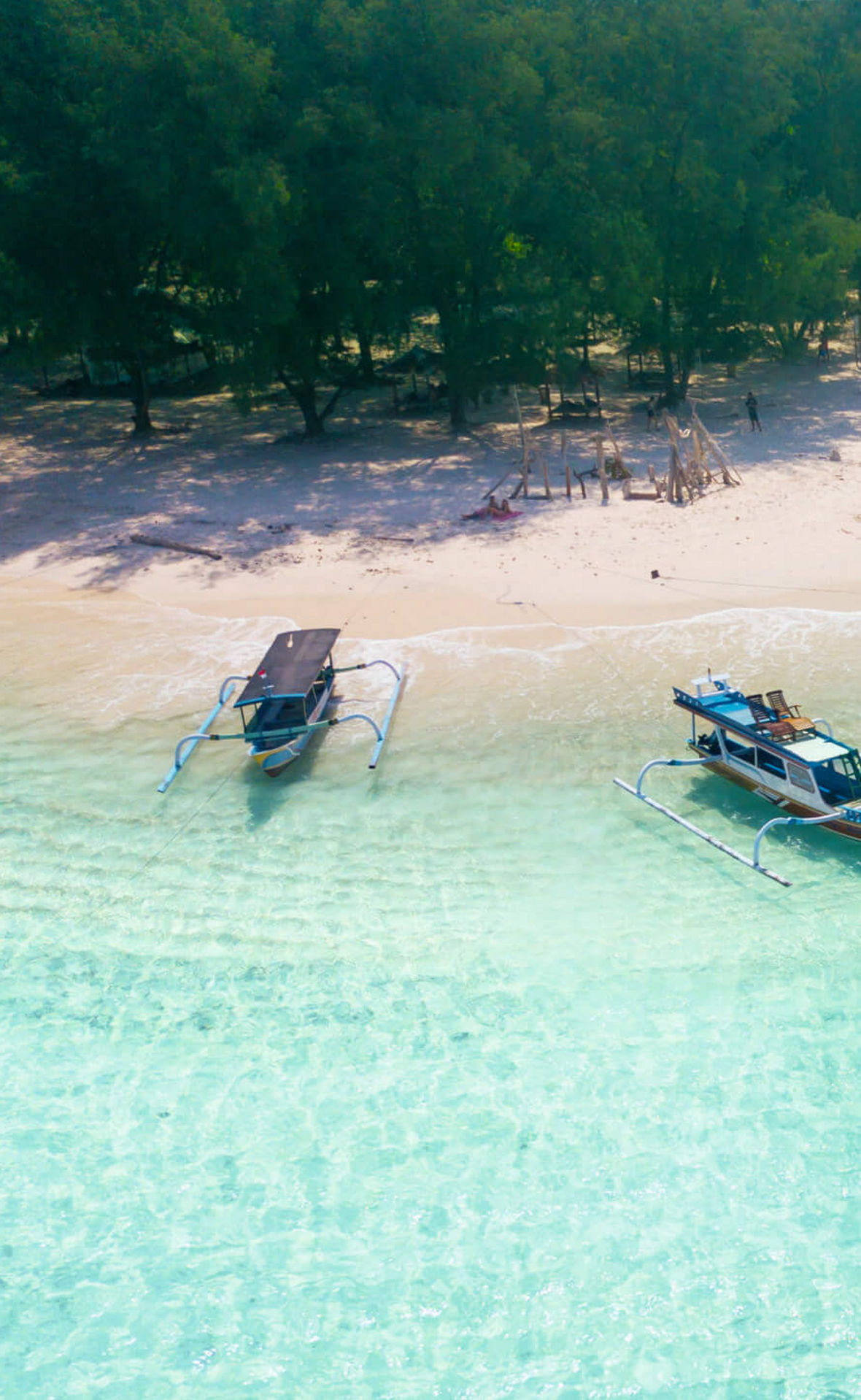 Ausflugsboote liegen an einem weißen Strand in kristallklarem Wasser. EInige Menschen sind am Strand, dahinter beginnt ein Wald.