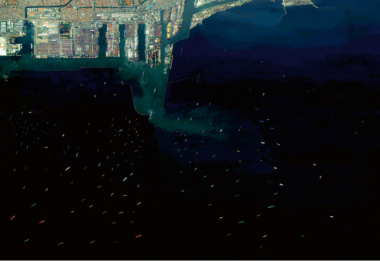 An aerial view of the port of Jakarta from a great height, with container ships moored in front of it waiting to enter.