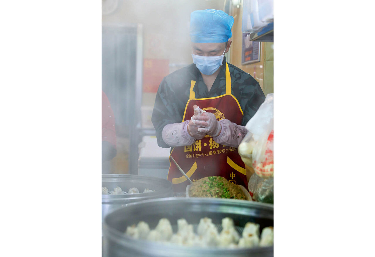 Ein Koch mit Haarschutz, Atemmaske und Schutzhandschuhen ind einer Garküche. Dampf steigt vor ihm auf.