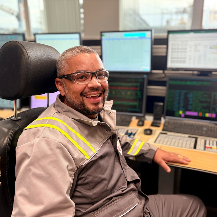 A man wearing glasses and a gray work uniform smiles while sitting in a control room chair.