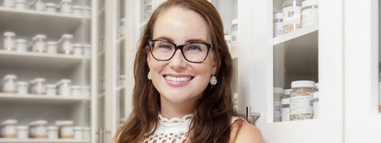 A woman stands confidently with arms crossed in front of shelves filled with jars and containers of various substances.