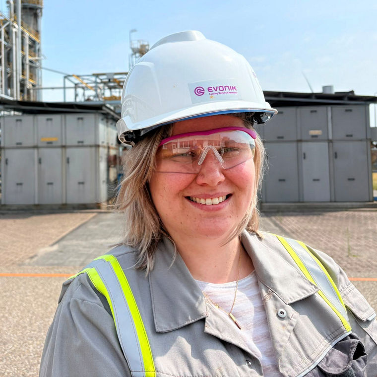 Woman wearing a white hard hat and safety glasses at an industrial plant on a sunny day.