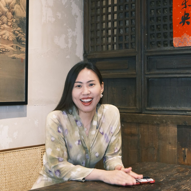 A woman in a patterned robe sits at a wooden table, holding a phone in a cozy, traditional setting.