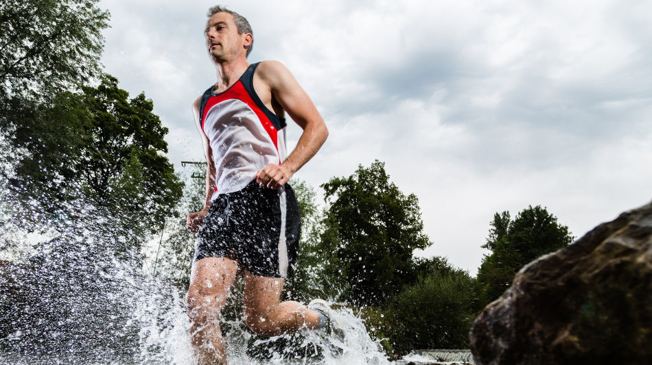 A man in athletic clothing running through water with splashes around him under a cloudy sky.