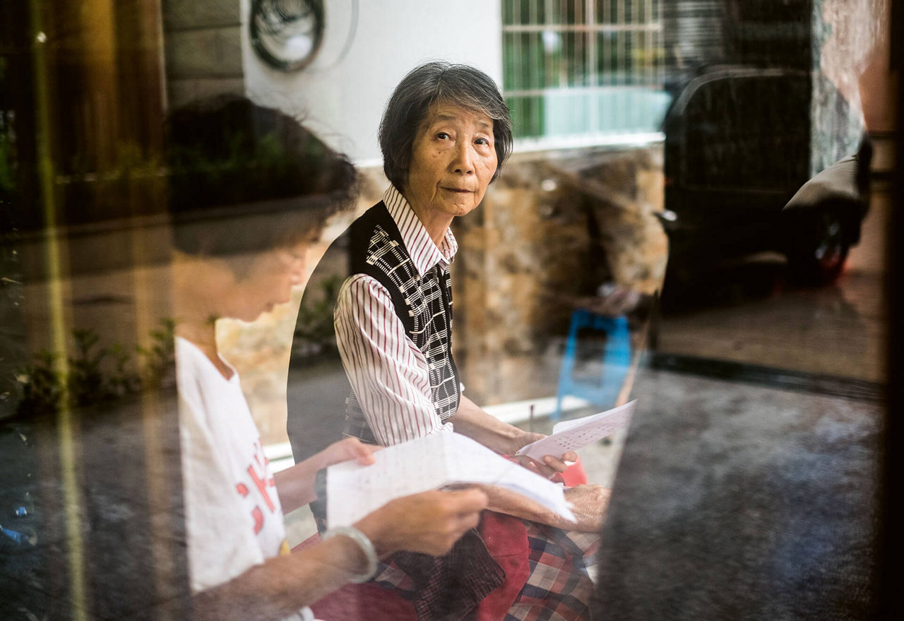 Eine Seniorin blickt durch eine Fensterscheibe in die Kamera. Neben ihr liest eine Frau etwas vor.