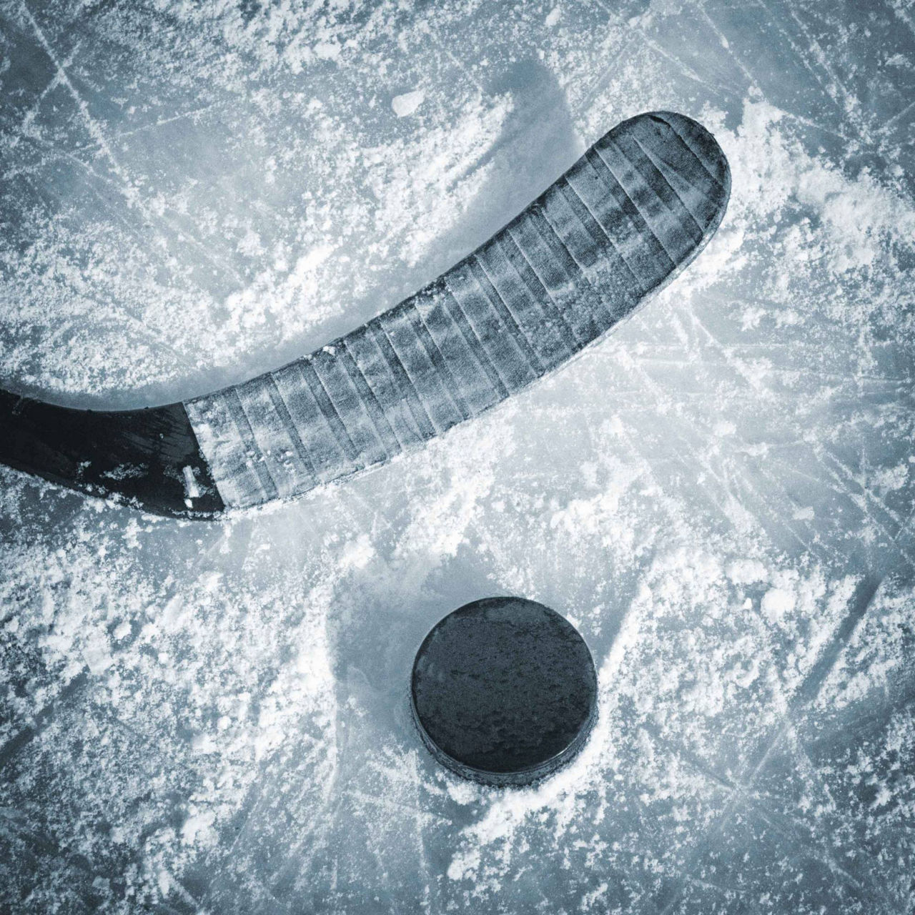 Close-up of a hockey stick blade and puck on a scratched, snow-dusted ice surface.