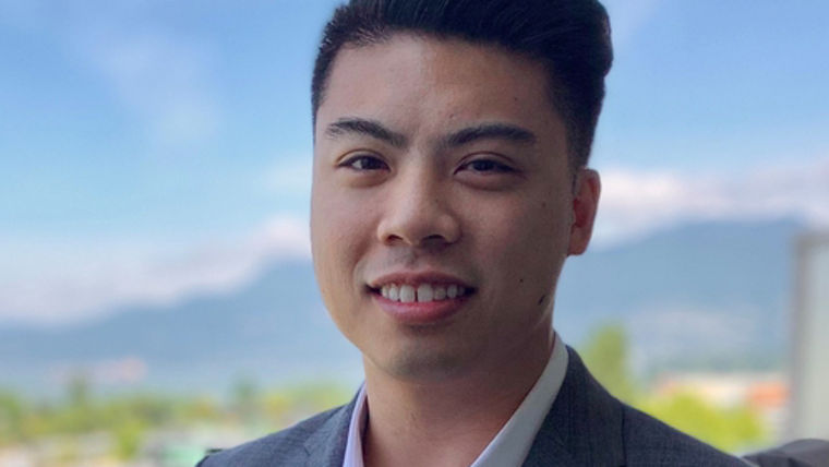 A man in a gray suit stands outdoors with a scenic backdrop of mountains and a clear sky.
