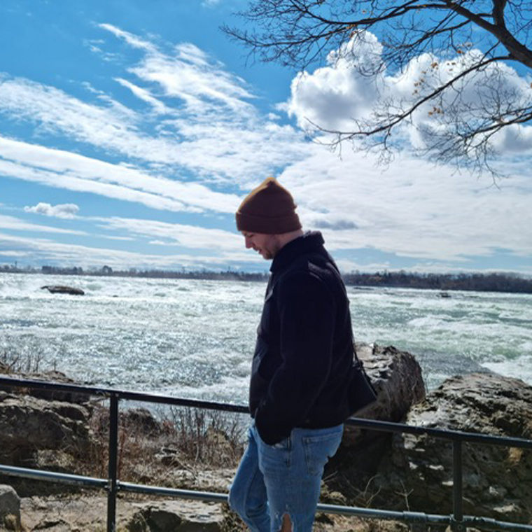 A person stands by a railing overlooking a turbulent river under a bright blue sky with clouds.