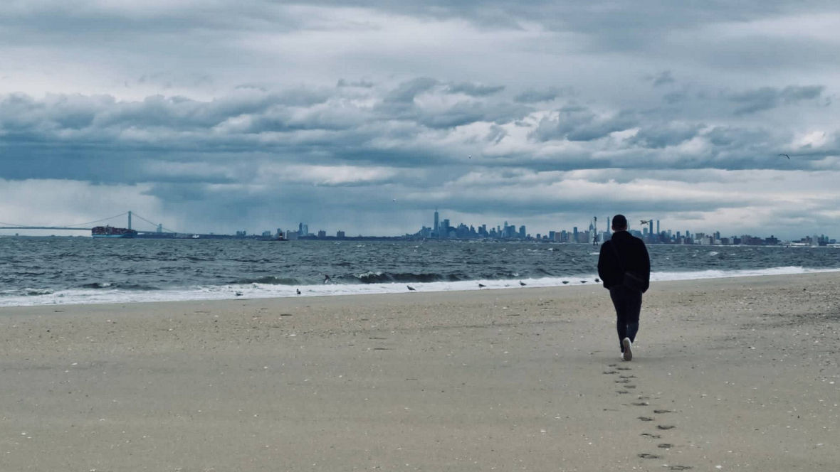 A person walks along a sandy beach with a skyline view in the background under a cloudy sky.
