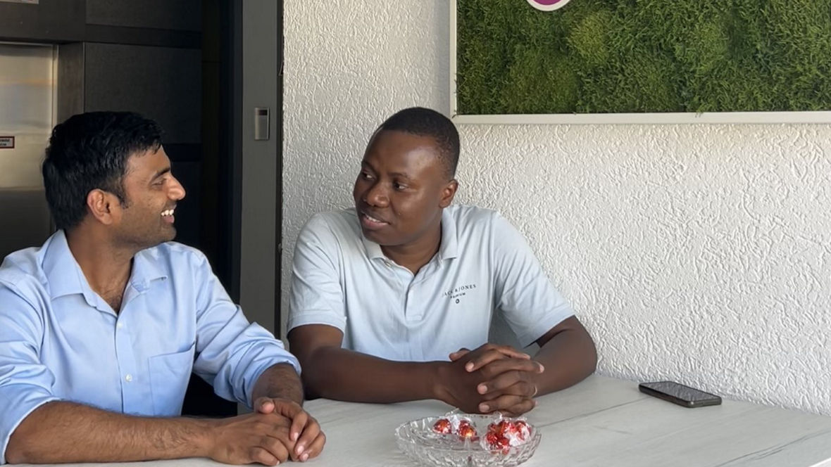 Two individuals are seated at a table in an office with an Evonik logo on the wall and a bowl of candies on the table.