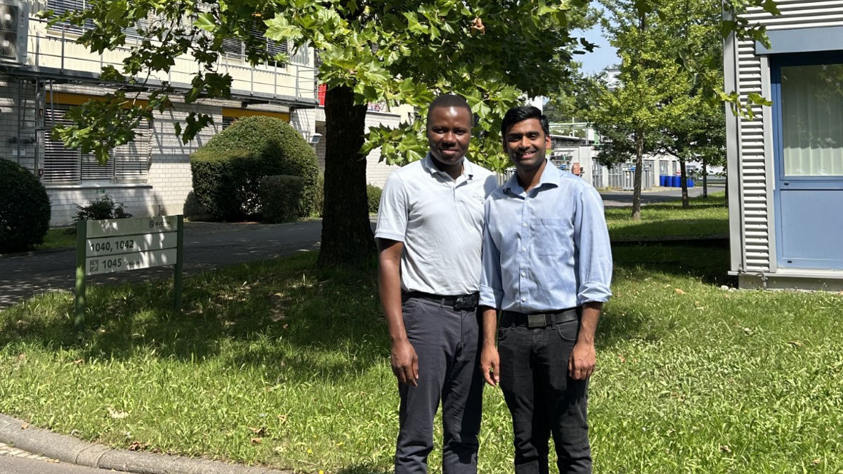 Two individuals stand together outside a building, surrounded by greenery and a sunny atmosphere.