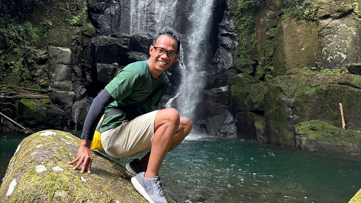 A person crouches on a mossy rock near a waterfall in a lush, green forest setting.