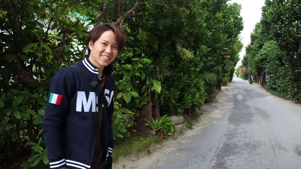 A person stands beside a lush, green pathway lined with trees, wearing a navy jacket with a logo.