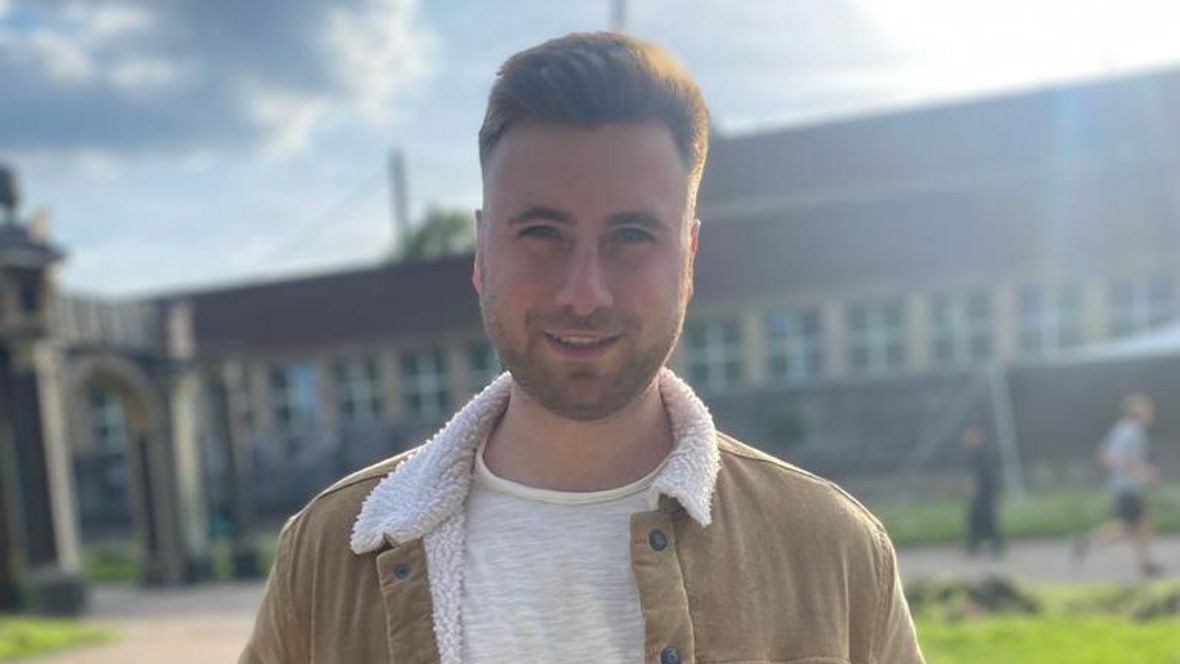 A person stands outdoors wearing a tan corduroy jacket with a white fleece collar and a light shirt, surrounded by greenery.