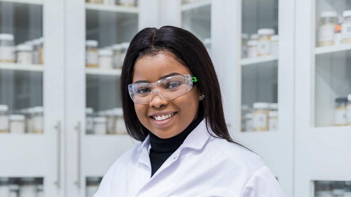 A woman in a white lab coat stands confidently in a laboratory filled with jars of materials.