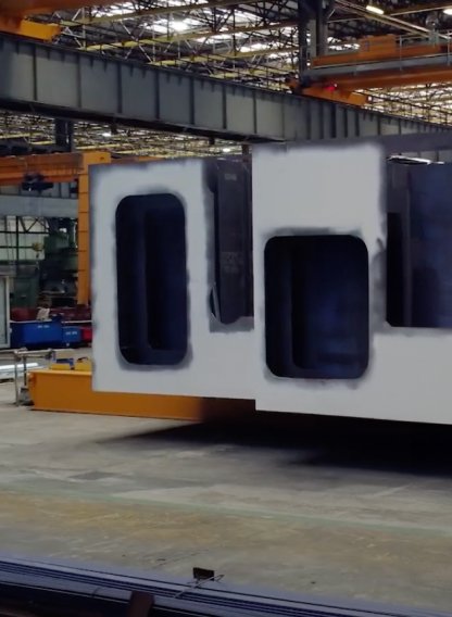 Fincantieri | Gruppo - Innovazione - Il nostro approccio all’innovazione: Grande componente rettangolare bianco con ampie aperture rettangolari e superficie lavorata, sostenuto da carrello elevatore industriale arancione, ambiente di fabbrica con travature, gru e macchinari sullo sfondo, illuminazione diffusa e pavimento in cemento usurato.