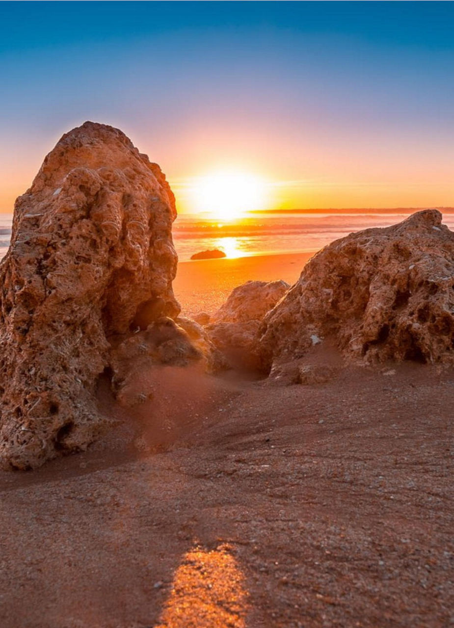 Gesteine im Vordergrund an einem Sandstrand bei Sonnenuntergang