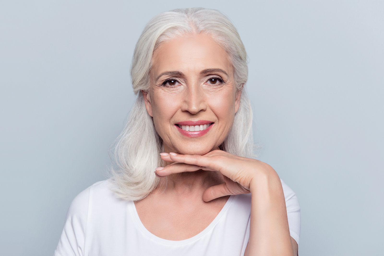 Charming old woman holding hand under chin with beaming smile looking at camera over gray background