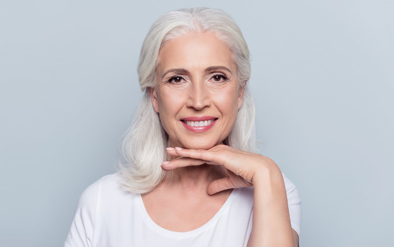 Charming old woman holding hand under chin with beaming smile looking at camera over gray background