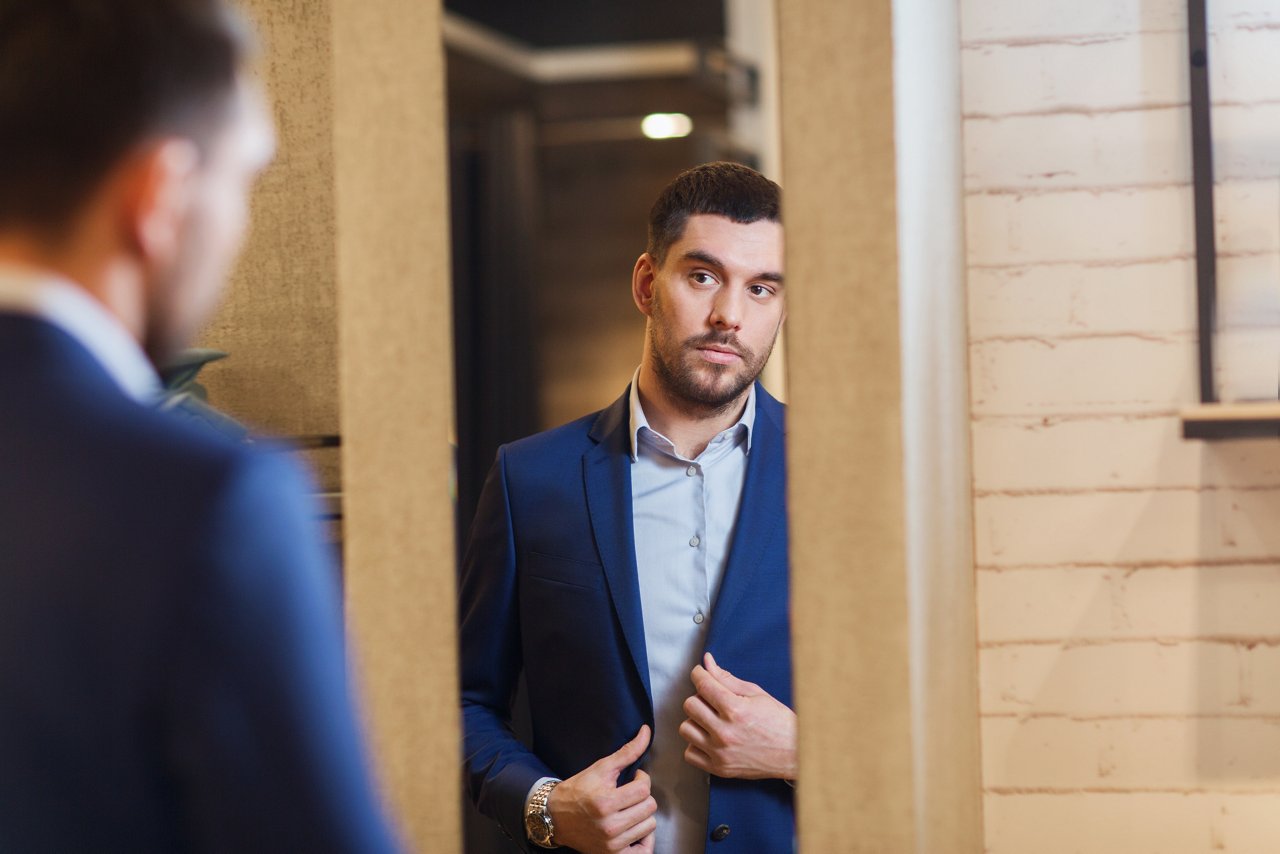 sale, shopping, fashion, style and people concept - elegant young man choosing and trying jacket on and looking to mirror in mall or clothing store