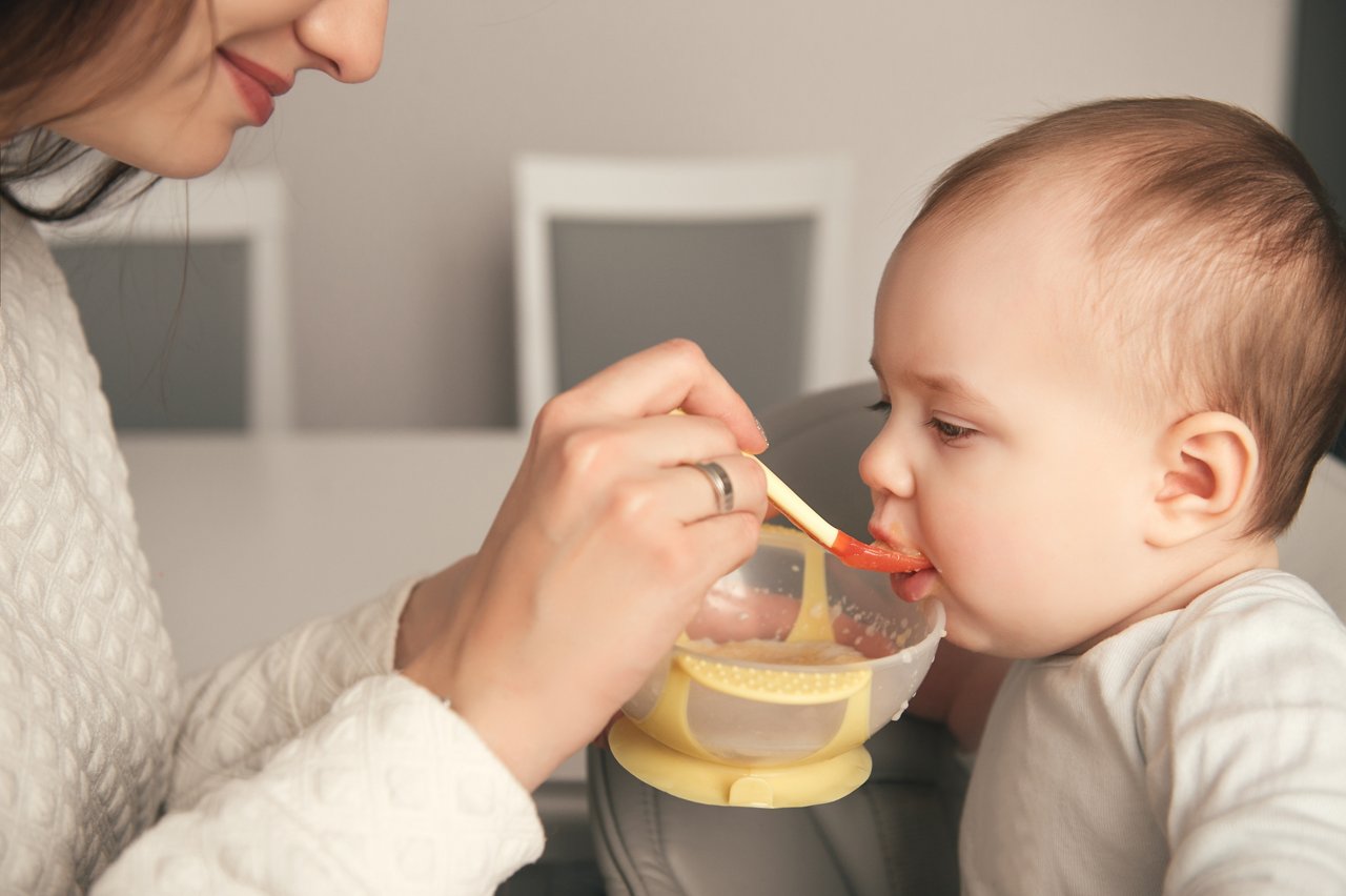 Mom feeding her baby girl with a spoon. Mother giving food to her eight-month child at home. Baby food. Close-up portrait.