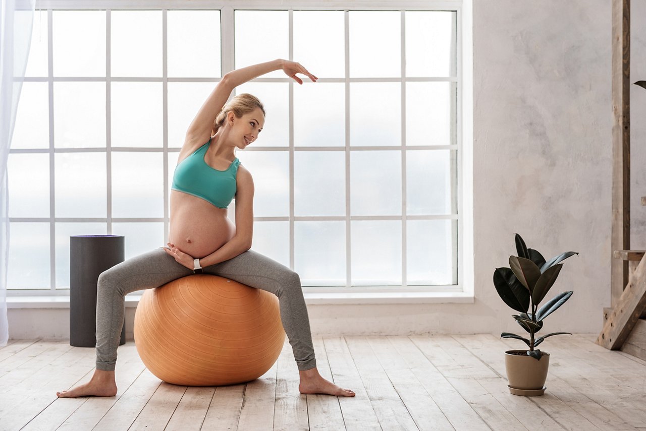 Strength of body and mind to be mother. Full length portrait of beautiful sporty pregnant woman making stretching fitness exercise on ball at home and smiling