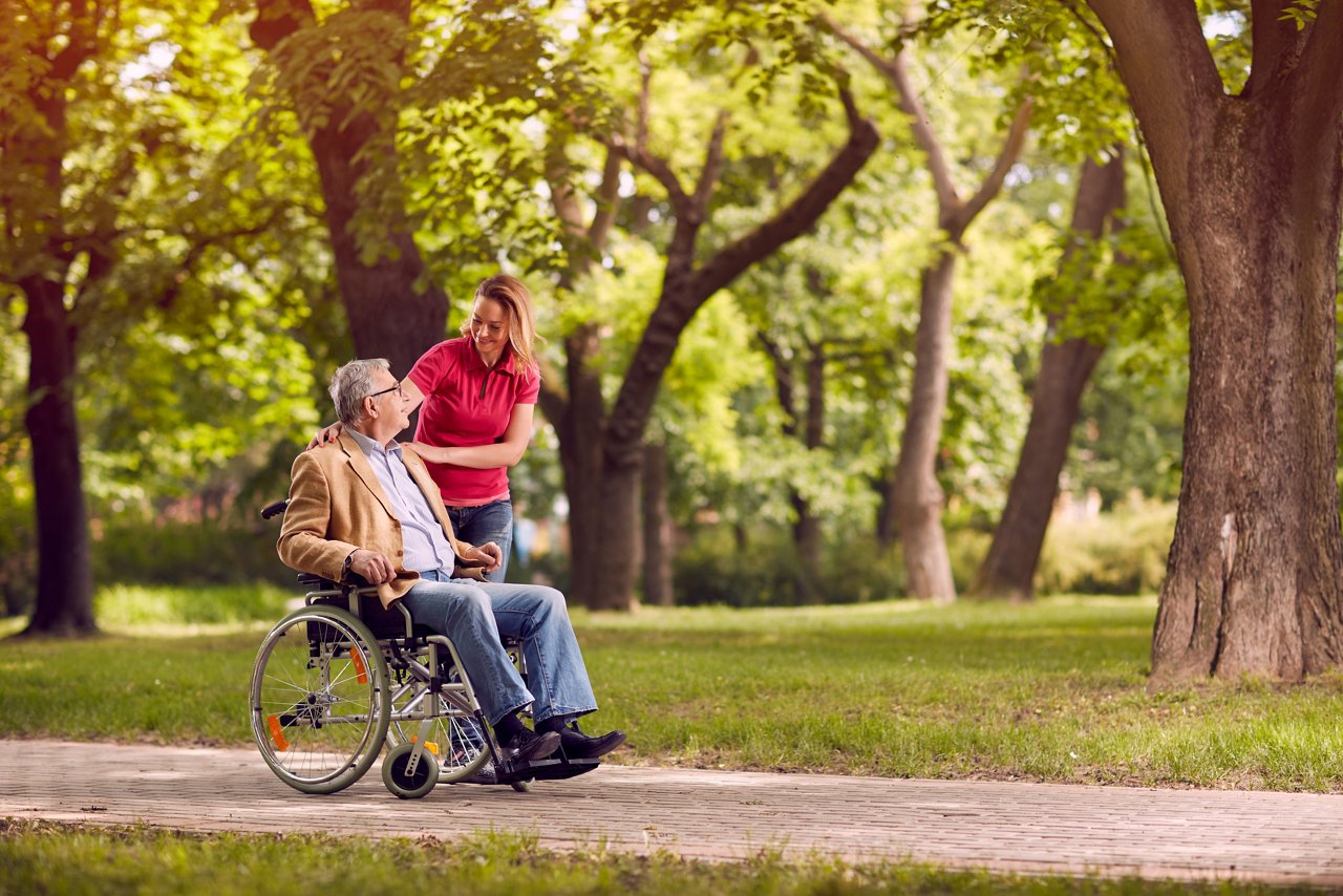 Happy senior man in wheelchair in the park with smiling daughter in the park
