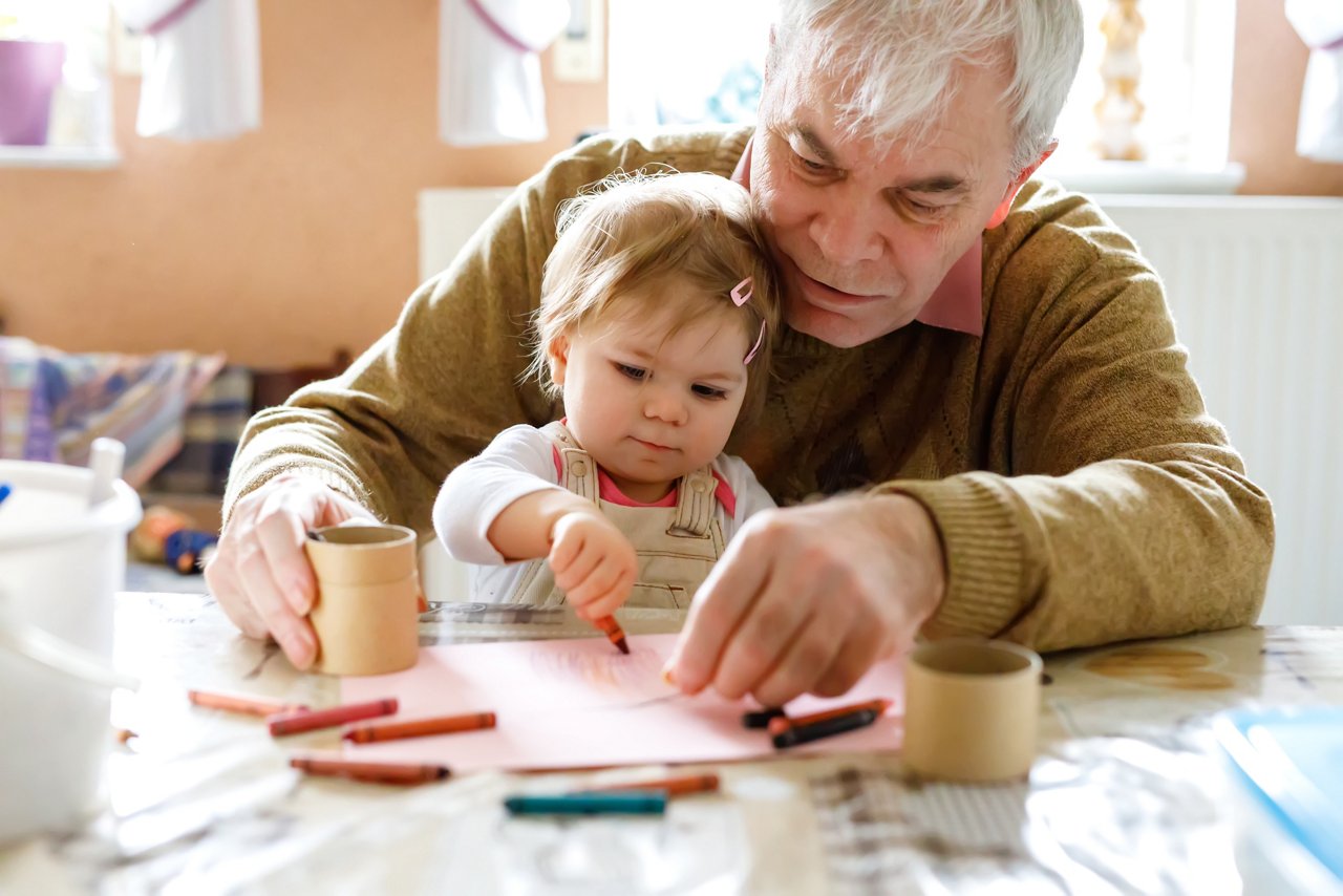 Cute little baby toddler girl and handsome senior grandfather painting with colorful pencils at home. Grandchild and man having fun together. Family and generation in love.