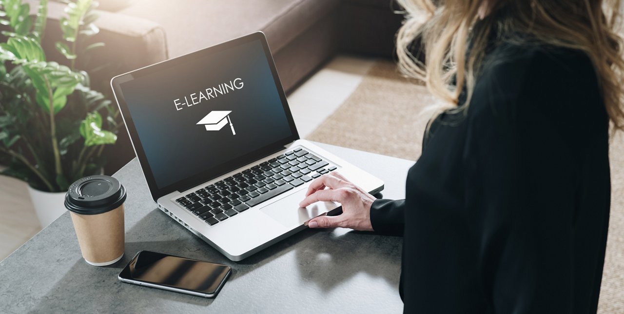 Frau steht am Tisch mit einem Laptop auf dem E-Learning steht, neben ihr ein Kaffeebecher und ein Handy