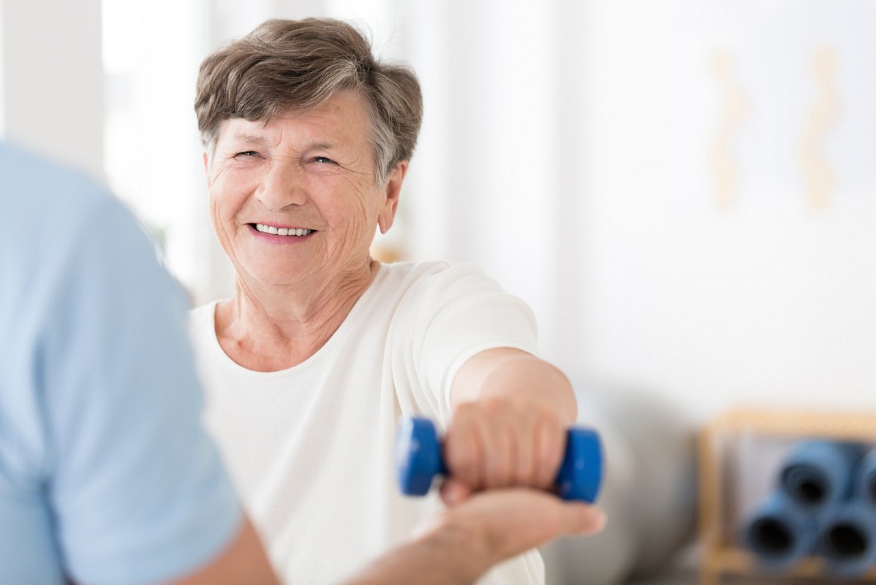 Seniorin während der Physiotherapie hält blaues Gewicht auf Schulterhöhe, Therapeut unterstützt während der Übung