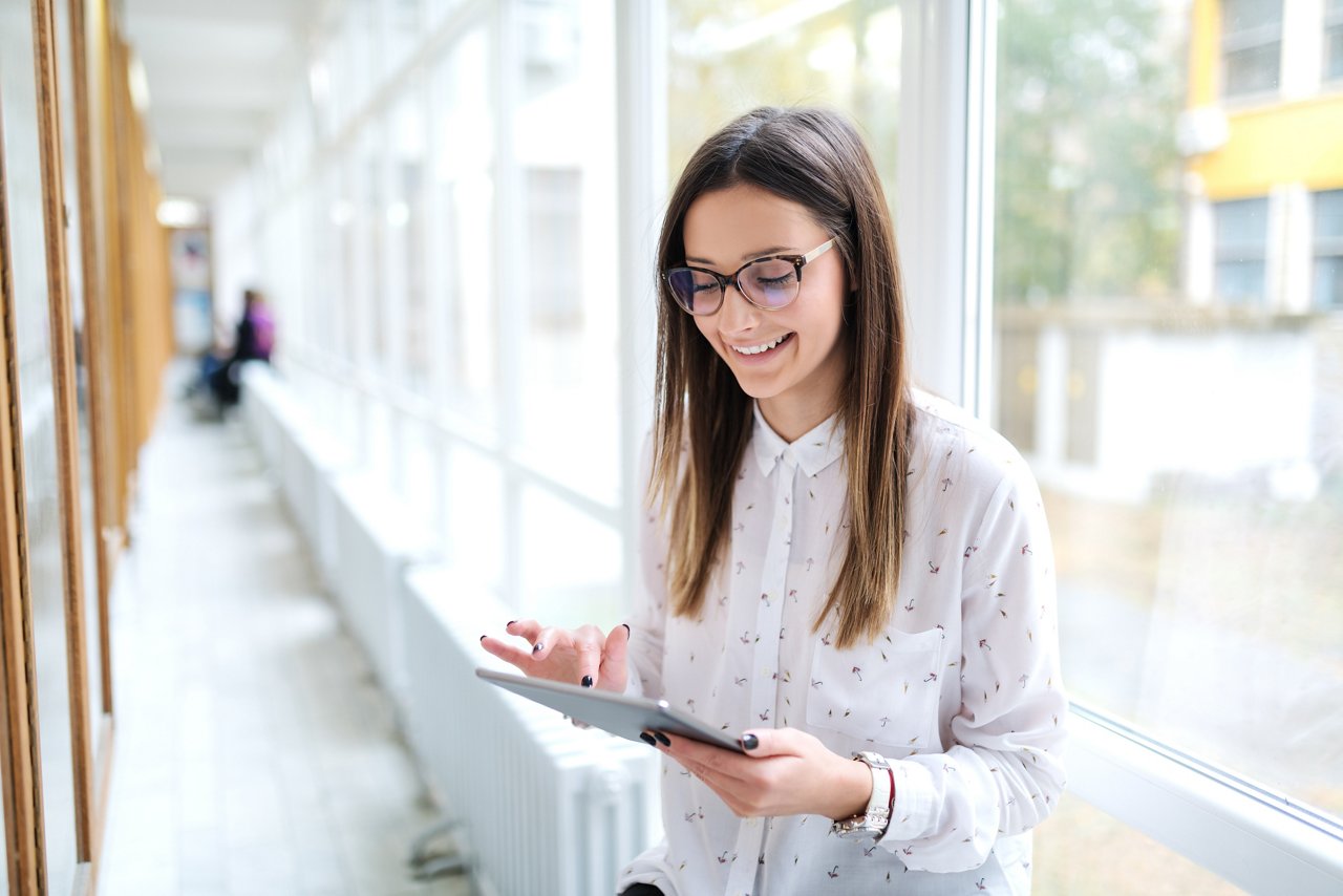 Close up of smiling Caucasian female student using tablet while standing next to noticeboard in college hall.